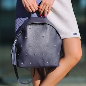May include: A dark blue leather backpack with silver studs and a tassel. The backpack has a zip closure and adjustable straps.