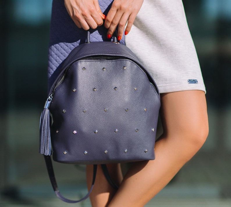 May include: A dark blue leather backpack with silver studs and a tassel. The backpack has a zip closure and adjustable straps.