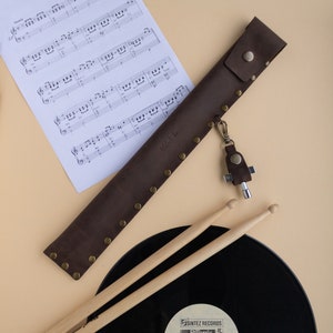 May include: A brown leather drumstick holder with a metal snap closure and a key chain with a multi-tool. The holder is lying on a beige surface next to a black vinyl record and two wooden drumsticks.