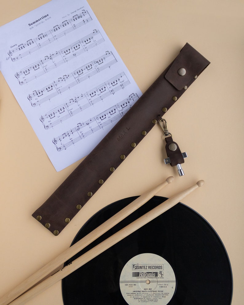 May include: A brown leather drumstick holder with a metal snap closure and a key chain with a multi-tool. The holder is lying on a beige surface next to a black vinyl record and two wooden drumsticks.