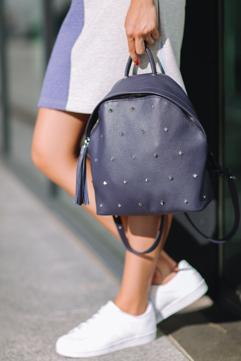 May include: A dark blue leather backpack with silver studs and a tassel. The backpack has a zipper closure and adjustable straps.