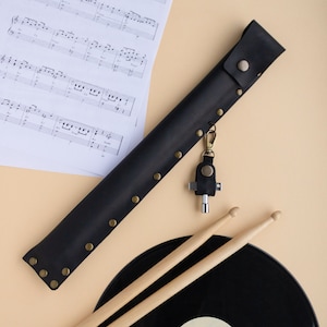May include: A black leather drumstick holder with a brass snap closure and rivets. The holder has a key chain attached with a small multi-tool. The holder is lying on a light brown surface next to a black vinyl record and two wooden drumsticks.