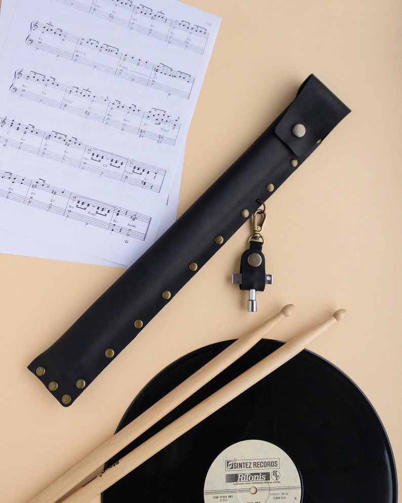 May include: A black leather drumstick holder with a brass snap closure and rivets. The holder has a key chain attached with a small multi-tool. The holder is lying on a light brown surface next to a black vinyl record and two wooden drumsticks.