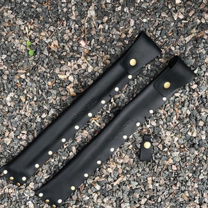 May include: Two black leather sheaths with gold studs and the text "ADAM & NOIR" and "ROMAN & NOIR" on them.
