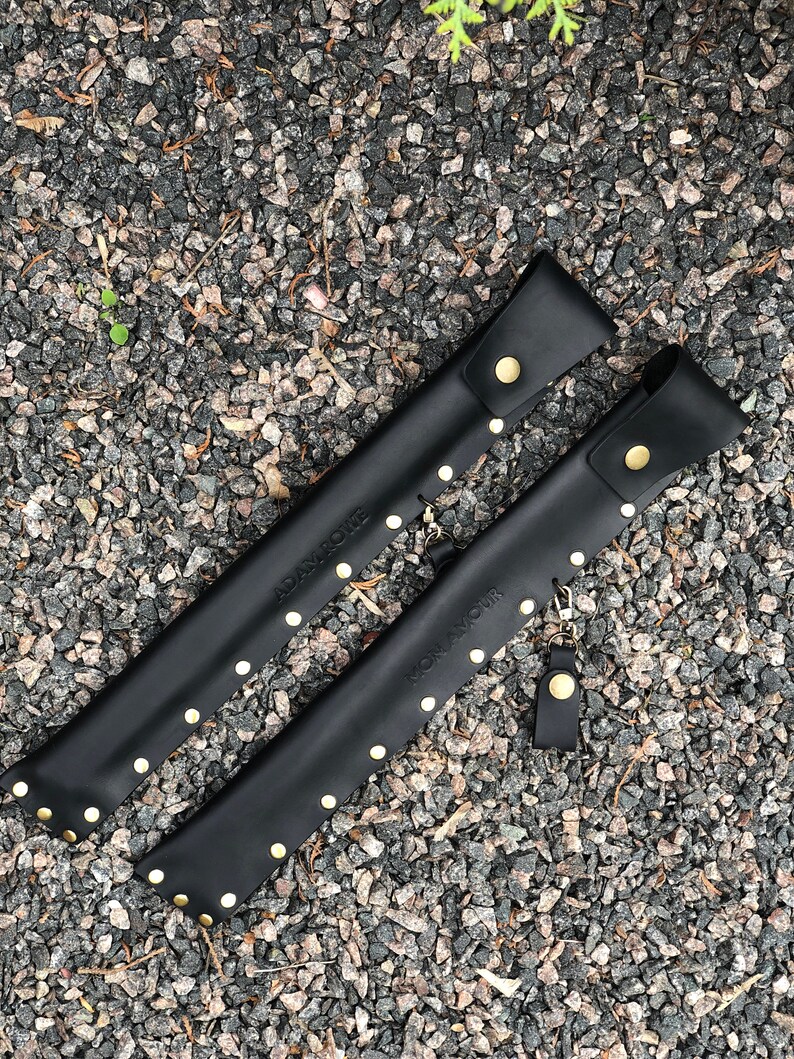 May include: Two black leather sheaths with gold studs and the text "ADAM & NOIR" and "ROMAN & NOIR" on them.