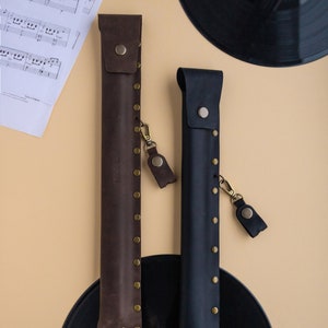 May include: Two leather cases for musical instruments, one brown and one black, with brass rivets and a small leather strap. The cases are on a black vinyl record.