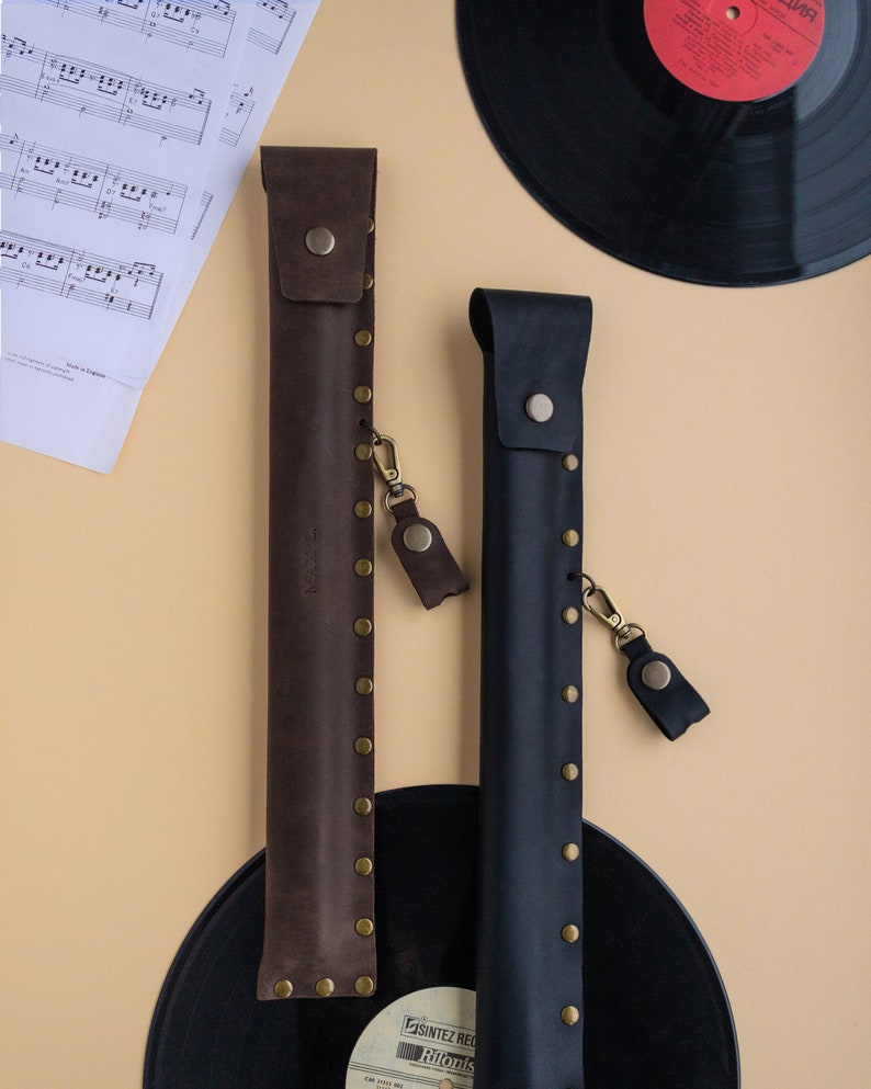 May include: Two leather cases for musical instruments, one brown and one black, with brass rivets and a small leather strap. The cases are on a black vinyl record.