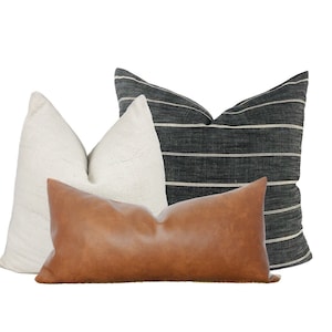 May include: Three decorative throw pillows. One is a long, rectangular brown leather pillow. The other two are textured, one is white and the other is dark gray with horizontal white stripes. All pillows are on a white background.