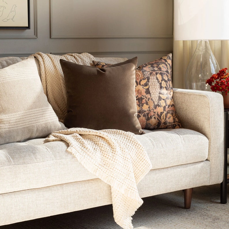 May include: A white couch with a brown throw blanket and three pillows. The pillows are a striped white and beige, a solid brown, and a patterned brown and beige.