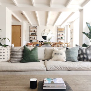 May include: A white couch with a variety of patterned throw pillows, including black and white, green, and blue. The couch is in a living room with a wooden coffee table and a large plant in the corner.