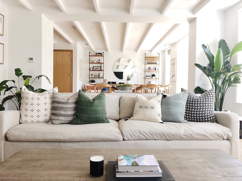 May include: A white couch with a variety of patterned throw pillows, including black and white, green, and blue. The couch is in a living room with a wooden coffee table and a large plant in the corner.