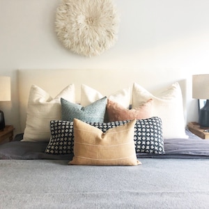 May include: A bed with a white headboard and a gray blanket. There are several pillows on the bed, including a tan pillow with stripes, a black and white patterned pillow, and a blue and green patterned pillow. There is a white feather wall decor above the headboard.