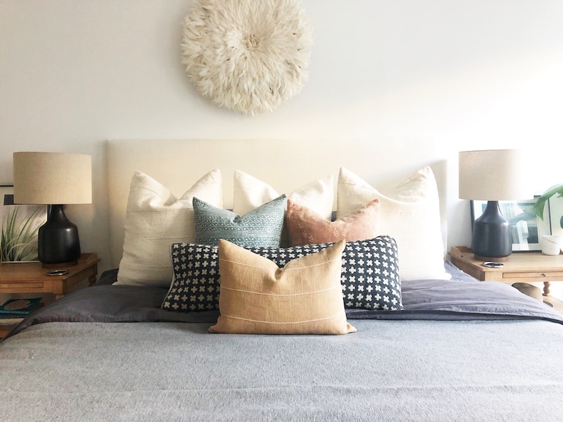 May include: A bed with a white headboard and a gray blanket. There are several pillows on the bed, including a tan pillow with stripes, a black and white patterned pillow, and a blue and green patterned pillow. There is a white feather wall decor above the headboard.
