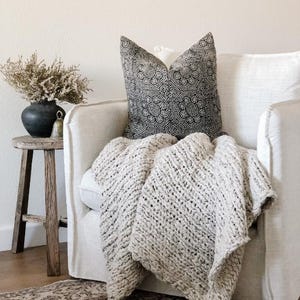 May include: A cozy living room scene featuring a cream-colored, chunky knit blanket draped over a white armchair. A decorative pillow with a black and white pattern rests on the chair. A small wooden side table holds a dark vase with dried flowers.