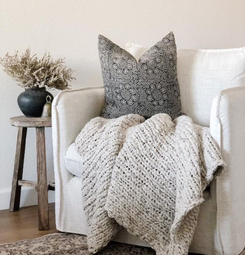 May include: A cozy living room scene featuring a cream-colored, chunky knit blanket draped over a white armchair. A decorative pillow with a black and white pattern rests on the chair. A small wooden side table holds a dark vase with dried flowers.