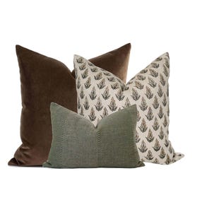May include: Four decorative cushions in a neutral colour palette. Two large brown velvet cushions flank a cushion with a cream background and a repeating black and brown leaf pattern. A smaller green textured cushion is in front.