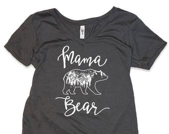 May include: A dark gray V-neck t-shirt with the text "Mama Bear" in white. A stylized white bear with a mountain scene inside is above the text.