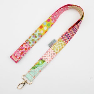 Könnte beinhalten: Ein farbenfrohes Patchwork-Lanyard mit einem goldenen Metallclip. Das Lanyard besteht aus verschiedenen Stoffen mit floralen, geometrischen und gepunkteten Mustern in den Farben Rosa, Gelb, Grün und Blau.
