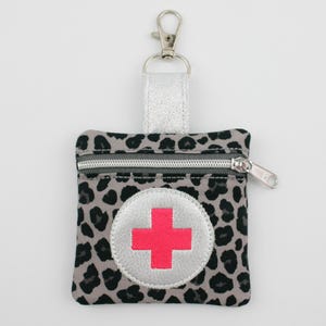 Può includere: Una pochette quadrata con cerniera e motivo leopardato grigio e nero. La parte anteriore presenta una toppa circolare argentata con una croce rosa brillante al centro. Un cinturino metallico argentato è fissato in alto con un moschettone.