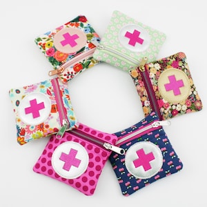Può includere: Una collezione di sei piccole pochette quadrate in tessuto disposte a cerchio. Ogni pochette presenta un diverso design floreale o fantasia, con un emblema a croce rosa su un cerchio bianco. Le pochette hanno chiusure con cerniera.