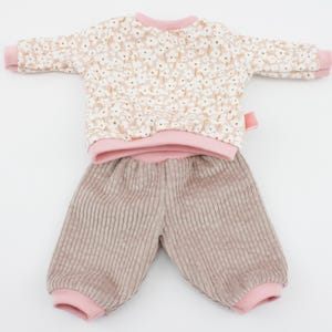 Può includere: Un completo per bambola con una maglia a maniche lunghe con stampa floreale e pantaloni di velluto a coste. La maglia ha scollatura e polsini rosa, e i pantaloni sono marrone chiaro con polsini rosa.