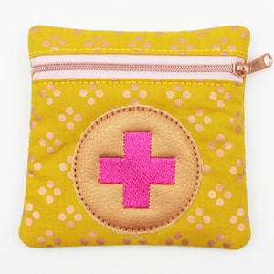 Può includere: Pochette gialla senape con cerniera e un motivo ripetuto di piccoli pois color rame. La pochette presenta una cerniera rosa e una toppa circolare con un disegno a croce rosa. La toppa è bordata con un materiale dorato.