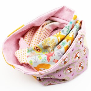 Könnte beinhalten: Ein farbenfrohes, wendbares Cowl-Schal mit verschiedenen Stoffmustern. Die Innenseite ist einfarbig rosa, während die Außenseite eine Mischung aus Designs wie Tupfen, Füchsen und floralen Motiven aufweist. Der Schal ist gefaltet.
