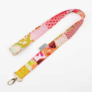 Könnte beinhalten: Ein farbenfrohes Patchwork-Lanyard mit einem goldenen Metallverschluss. Das Lanyard besteht aus verschiedenen Stoffen mit floralen, gepunkteten und geometrischen Mustern in Rosa-, Rot- und Gelbtönen.