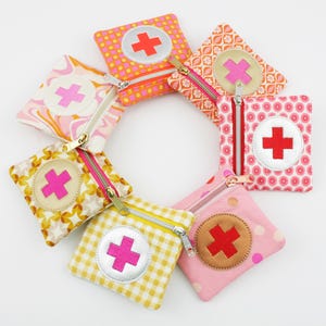Può includere: Una collezione di piccole pochette con cerniera in vari modelli e colori, disposte a forma circolare. Ogni pochette presenta un simbolo a croce, con colori tra cui rosa, rosso e arancione. Le pochette hanno cerniere color oro.