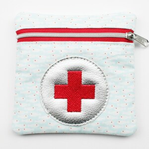 Könnte beinhalten: Eine kleine Tasche aus blauem und weißem Stoff mit einem roten Kreuz, das auf einen silbernen Kreis gestickt ist. Die Tasche hat einen Reißverschluss.