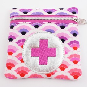 Può includere: Una pochette quadrata in tessuto con un motivo a squame ripetuto nei toni del rosa, viola e rosso. La pochette presenta una chiusura a zip argentata e una toppa circolare argentata al centro, con una croce ricamata in rosa brillante.