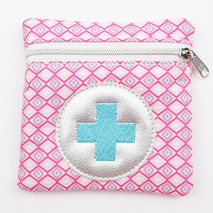 Può includere: Una piccola pochette rosa con cerniera con un motivo geometrico bianco e rosa. Un cerchio argentato con un segno più blu è centrato sulla pochette.