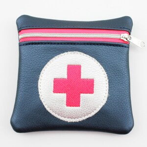 Könnte beinhalten: Eine marineblaue Tasche mit rotem Reißverschluss und einem weißen und rosa Kreuzemblem. Die Tasche ist quadratisch und hat einen silbernen Reißverschluss. Das Kreuz ist mittig auf der Vorderseite.