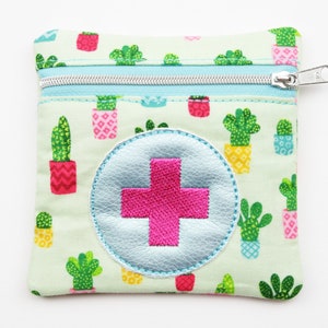 Könnte beinhalten: Eine kleine, grüne Reißverschlusstasche mit weißem Hintergrund und einem sich wiederholenden Muster aus Topfkakteen. Die Tasche hat einen großen, blauen Kreis mit einem rosa Pluszeichen in der Mitte. Die Tasche ist perfekt zum Aufbewahren von kleinen Gegenständen wie Medikamenten oder Erste-Hilfe-Utensilien.