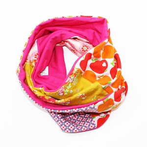 Könnte beinhalten: Ein farbenfroher Loop-Schal mit Patchwork-Design. Der Schal hat eine Mischung aus Mustern, darunter Blumen, Punkte und herzförmige Früchte. Die Innenseite ist einfarbig leuchtend pink, und der Schal ist aus weichem Stoff.