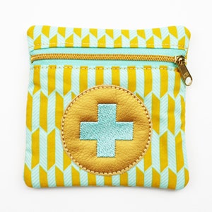 Può includere: Piccola pochette quadrata con cerniera dorata e motivo geometrico giallo e azzurro. Un cerchio in pelle dorata con una croce azzurra è centrato sul davanti. La pochette è probabilmente per trasportare piccoli oggetti.