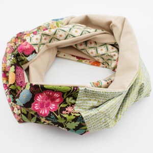 Könnte beinhalten: Ein farbenfroher Loop-Schal mit Blumenmustern in Rosa, Blau und Orange auf dunklem Braun. Der Schal hat auch ein beiges Innenfutter und Abschnitte mit grünen und weißen geometrischen Designs. Ein stilvolles Accessoire.