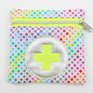 Può includere: Una piccola pochette con cerniera colorata con sfondo bianco e un cerchio argentato con un segno più giallo neon al centro. La pochette è realizzata in tessuto con un motivo ripetitivo di piccoli cerchi e croci.