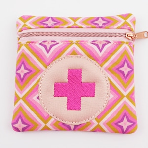 Può includere: Un astuccio quadrato con cerniera e motivo geometrico rosa, giallo e bianco. Una croce rosa è ricamata su un cerchio rosa chiaro sul davanti. La cerniera e il tirante sono in oro rosa.