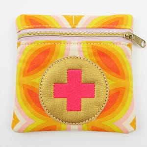 Può includere: Una piccola pochette con cerniera gialla, arancione e rosa con un cerchio dorato e un segno più rosa al centro. La pochette è realizzata in tessuto con un motivo geometrico.