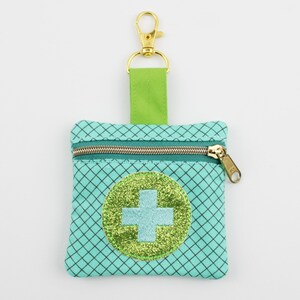Può includere: Una pochette quadrata color verde menta con cerniera, caratterizzata da un motivo a griglia nera e una toppa circolare centrale con sfondo verde lime glitterato e una croce azzurra. Include una linguetta in tessuto verde lime e un moschettone dorato.