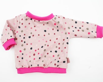 Puede incluir: Sudadera rosa claro para bebé con un estampado de lunares coloridos. La parte superior de manga larga tiene puños, cuello y dobladillo de canalé rosa brillante. Una pequeña etiqueta rosa está cosida en el lateral.