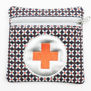 Könnte beinhalten: Eine kleine Reißverschlusstasche mit einem weißen und blauen Blumenmuster. Die Tasche hat einen silbernen Reißverschluss und einen silbernen Kreis mit einem roten Kreuz in der Mitte.