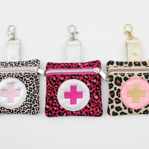 Può includere: Tre pochette quadrate con cerniera e stampa leopardata. Ogni pochette presenta una toppa circolare con un simbolo a croce al centro e un moschettone in metallo argentato o dorato fissato a un passante in tessuto per appenderla.