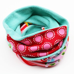 Könnte beinhalten: Ein farbenfroher Halsschmeichler mit türkisfarbenem Fleecefutter und einer Außenseite aus gemustertem Stoff. Die Außenseite hat roten Stoff mit rosa Punkten, gestreiften Stoff und Blumenstoff. Der Halsschmeichler ist zum Tragen um den Hals konzipiert.