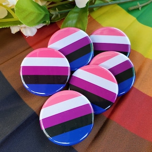 Puede incluir: Seis botones redondos con los colores de la bandera del orgullo bisexual: rosa, morado, azul y negro. Los botones están dispuestos en círculo sobre un fondo de tela arcoíris.