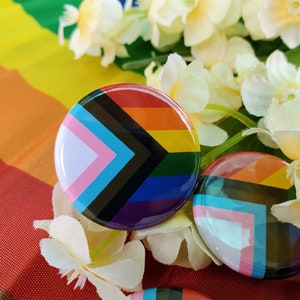 May include: Close-up of a set of colorful button pins featuring a rainbow flag design with a black and brown chevron. The pins are surrounded by white flowers.