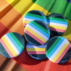 Op de afbeelding: Zes ronde, zwart-wit gestreepte knoppen met groene, blauwe en roze strepen. De knoppen zijn in een halve cirkel op een regenboogkleurige stoffen achtergrond gerangschikt.