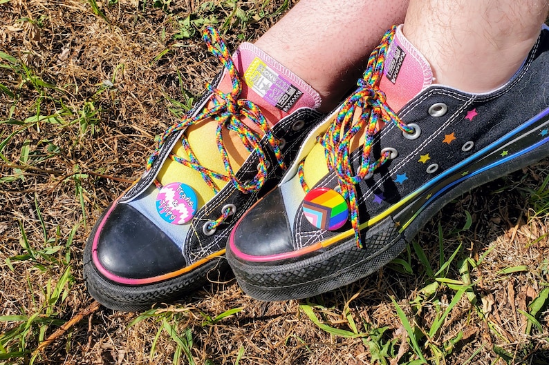 Puede incluir: Un par de zapatos de lona negros con cordones arco&iacute;ris y una franja arco&iacute;ris a lo largo de la suela. Un zapato tiene un pin que dice "Trans & Valid" y el otro tiene un pin con un arco&iacute;ris y un s&iacute;mbolo transg&eacute;nero.