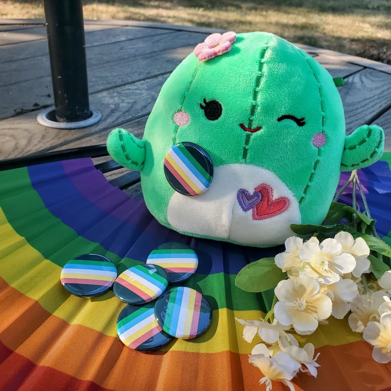 Puede incluir: Un juguete de peluche de cactus verde con una flor rosa en la parte superior y una cara sonriente. El cactus sostiene un coraz&oacute;n arco&iacute;ris y est&aacute; rodeado de botones de presi&oacute;n arco&iacute;ris.